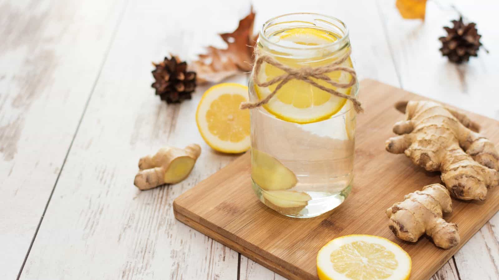 A mason jar filled with lemon-infused water sits on a wooden board. Lemon slices float inside, while more slices and ginger roots are placed nearby. Pine cones and a dried leaf rest in the background on a light wooden surface.