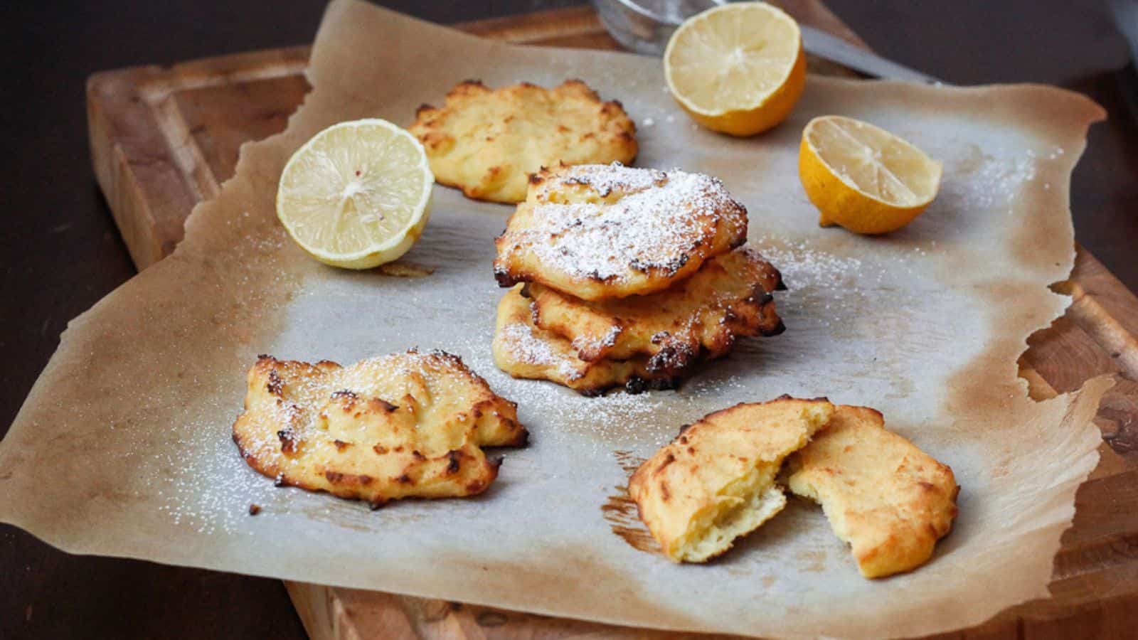 Four lemon ricotta cookies with powdered sugar are on a parchment-lined wooden board. A halved lemon is nearby, adding a fresh and zesty touch to the scene. One cookie is broken in half, displaying its soft interior.