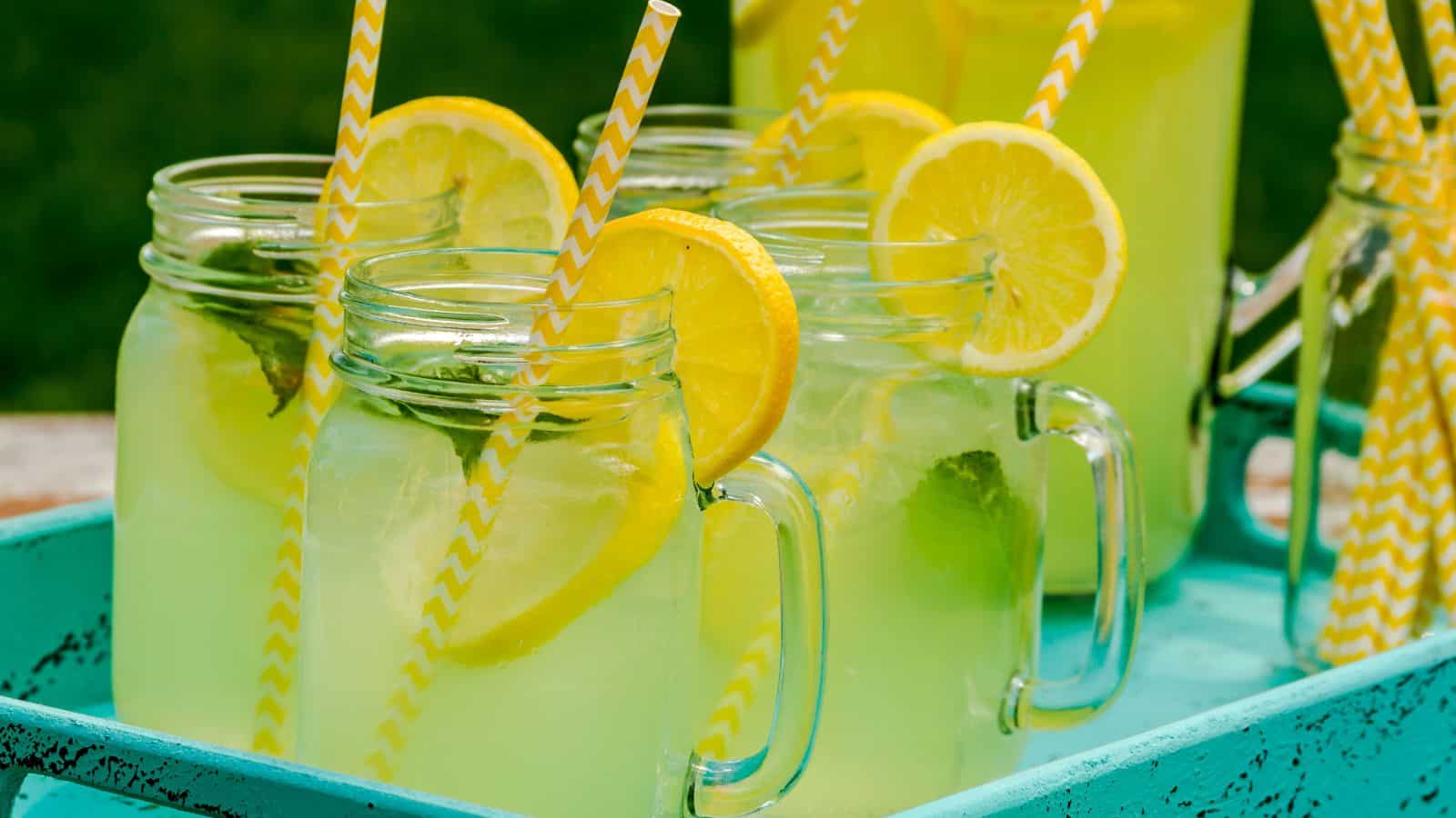 Mason jars filled with lemonade sit on a turquoise tray. Each jar is garnished with lemon slices and has a yellow and white striped straw.