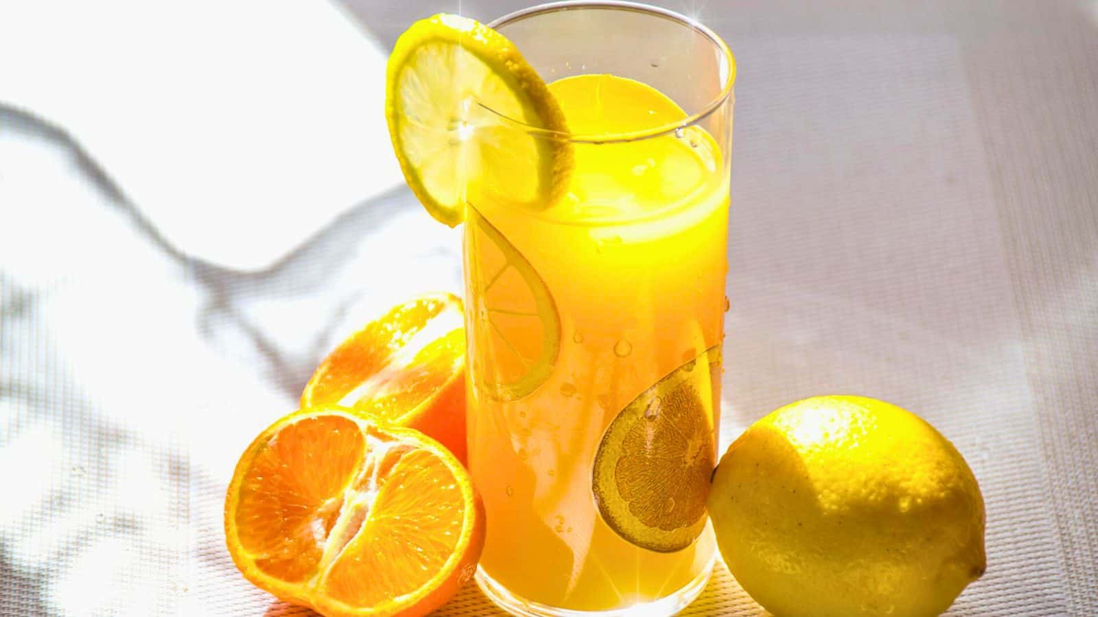 A glass of orange juice with a lemon slice on the rim sits on a white surface. There are two orange halves and a whole lemon nearby, with bright lighting casting soft shadows.