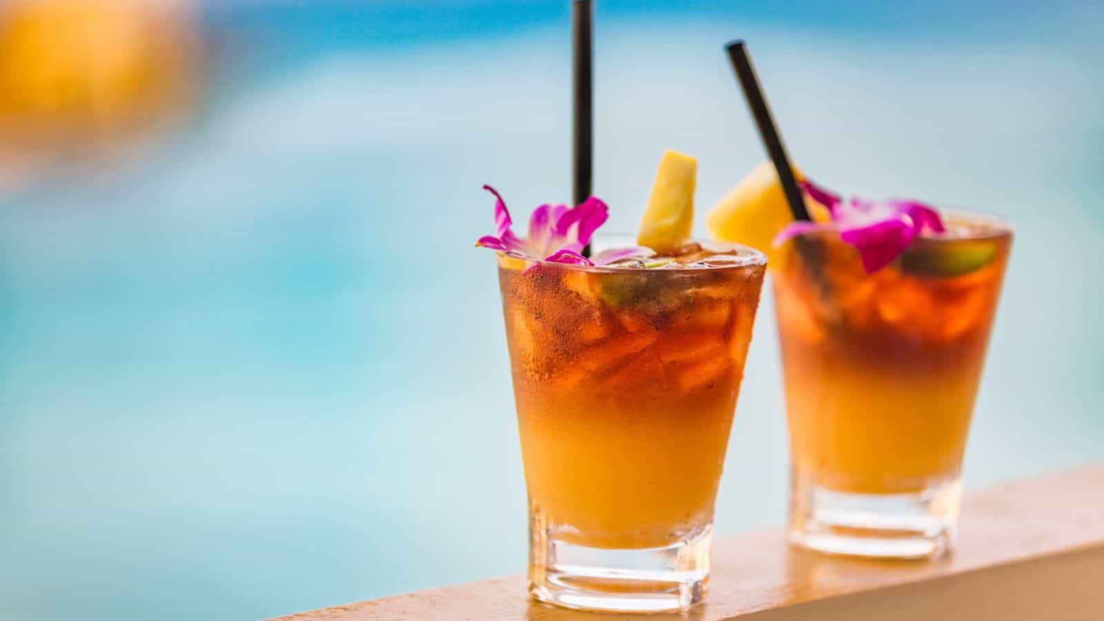 Two tropical cocktails in clear glasses sit on a ledge against a blurred blue background. Each drink is garnished with a slice of pineapple, a purple orchid, and a black straw. The drinks have a gradient from orange to dark brown.