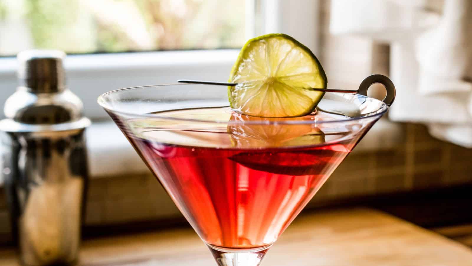 A close-up of a martini glass filled with a red cocktail, garnished with a lime wheel on a cocktail pick. A cocktail shaker is blurred in the background, sitting on a kitchen countertop with soft natural light coming in through a window.