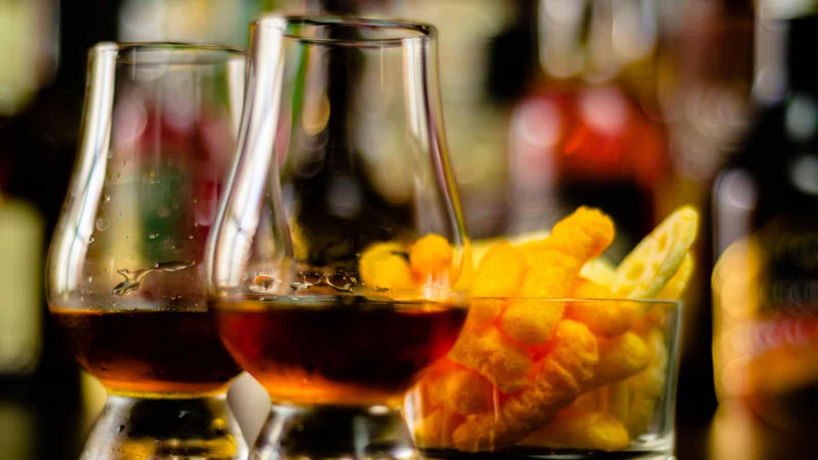 Two glasses containing a dark liquid are placed next to a small glass bowl filled with cheese puffs. The background is blurred, featuring various indistinct bottles. The scene suggests a casual or social setting.