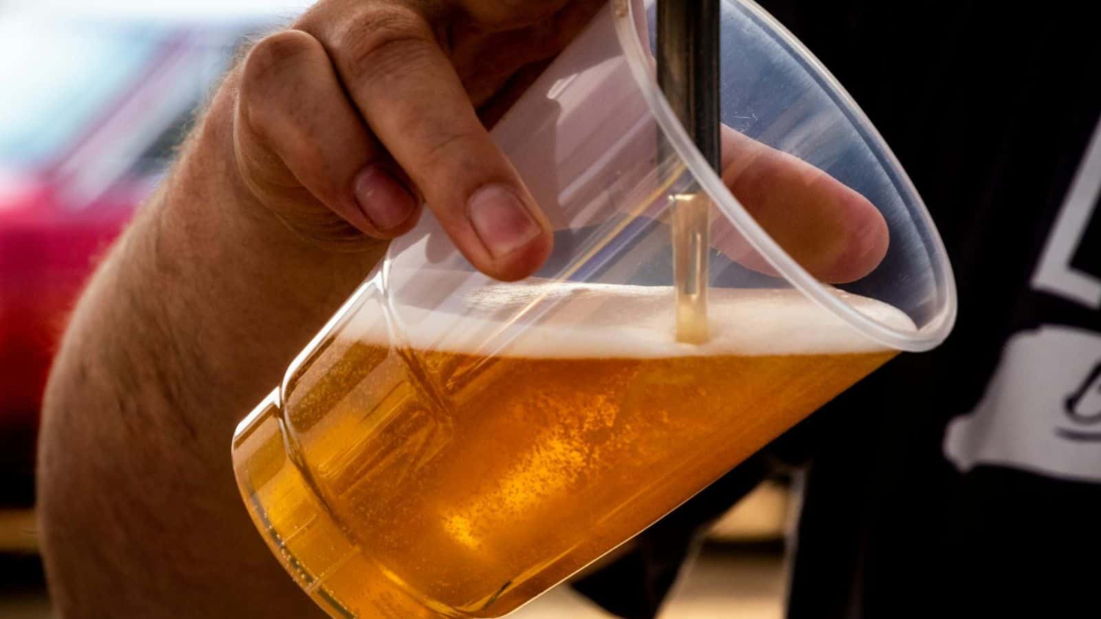 A person pours beer from a tap into a plastic cup, tilting it to reduce foam. The beer is golden with a frothy head forming at the top. The background is softly blurred, highlighting the focus on the action of pouring.