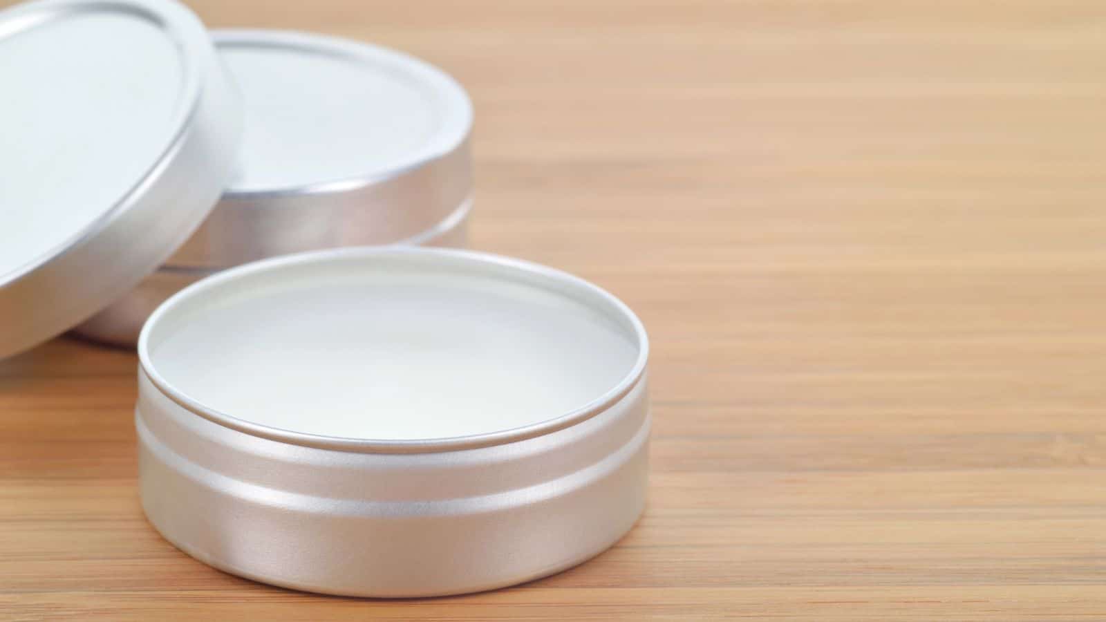 An open metal tin containing a white solid substance is placed on a wooden surface. Another similar tin with the lid partially open is in the background.