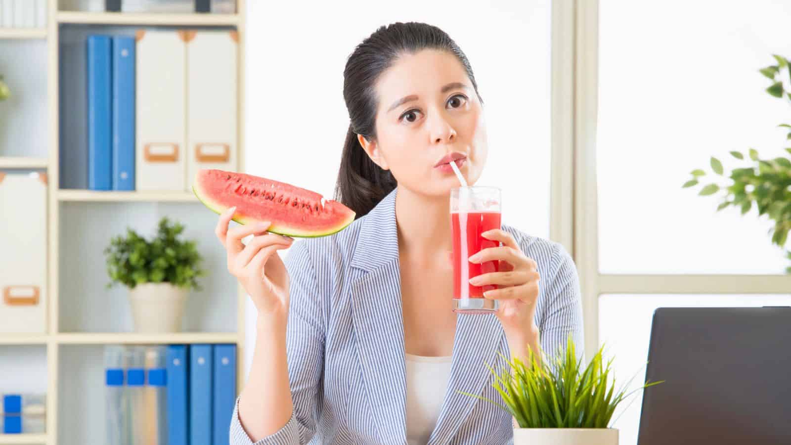 A person in a blue pinstripe blazer is sitting at a desk, holding a slice of watermelon and drinking watermelon juice through a straw. In the background, there are shelves with books and plants, and a laptop is visible on the desk.