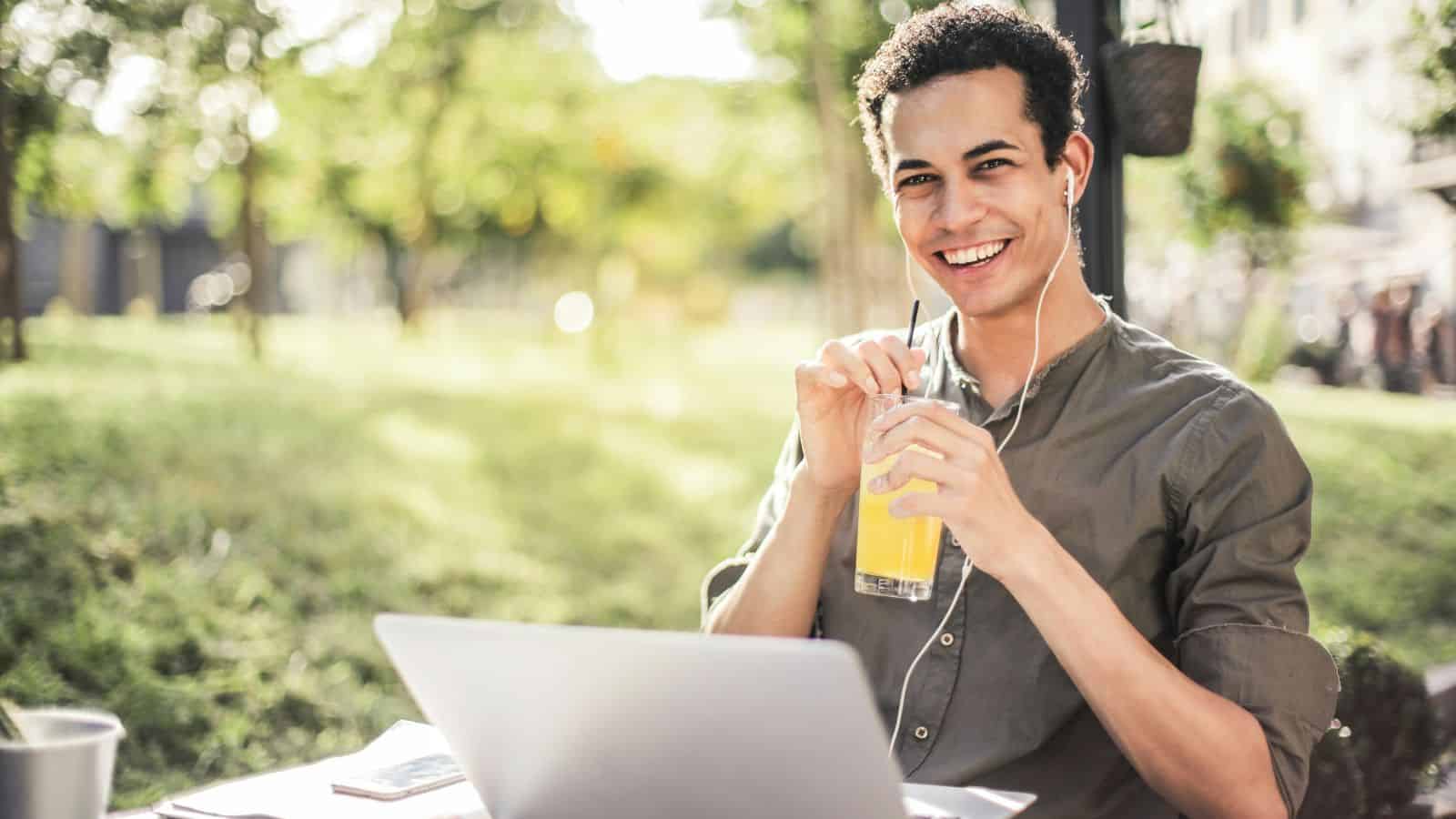 A person sits outdoors at a table with a laptop, holding a glass of orange juice and smiling. They are wearing earphones and a casual green shirt. The background features greenery and soft sunlight.