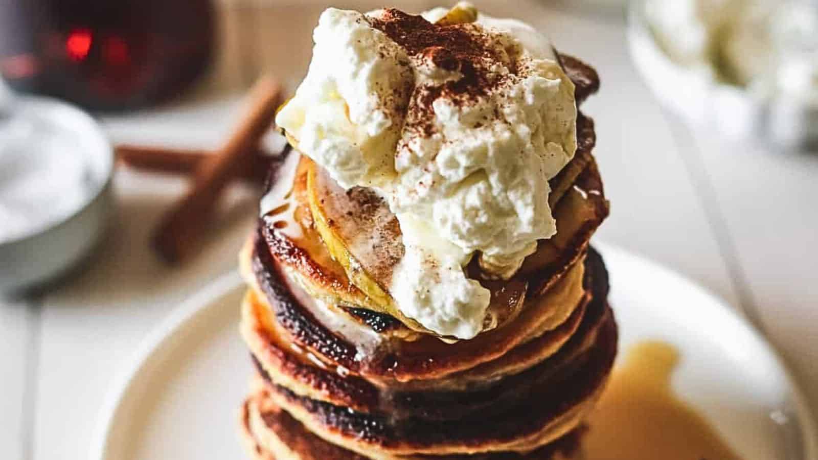 A stack of pancakes topped with whipped cream and a sprinkle of cinnamon on a white plate. Syrup is drizzling down the sides, and a cinnamon stick is visible in the background.