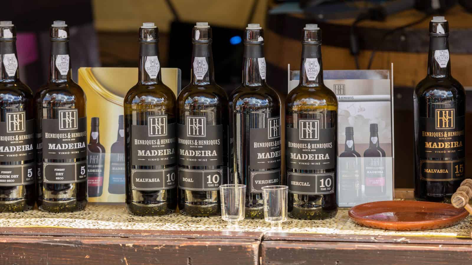 A display of seven bottles of Madeira wine with labels indicating different varieties, including Malvasia, Sercial, and Verdelho. Two empty wine glasses are in front, and promotional materials are visible in the background.