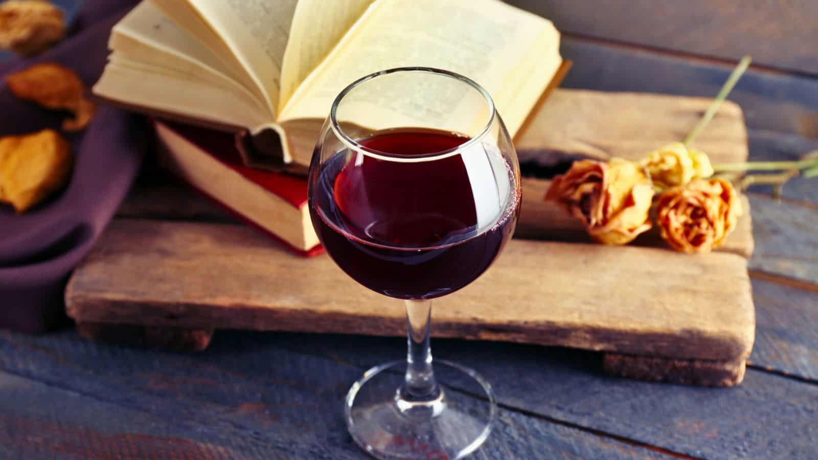 A glass of red wine sits on a rustic wooden surface next to an open book and a closed book. Two dried roses are also present, adding a touch of elegance to the scene.