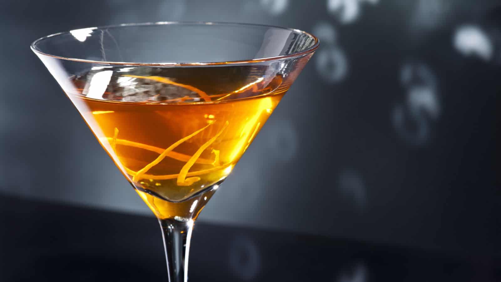 A close-up of a martini glass filled with an amber-colored liquid, garnished with a twist of lemon peel. The background is dark with soft, out-of-focus circular lights. The glass is positioned to the left side of the image.