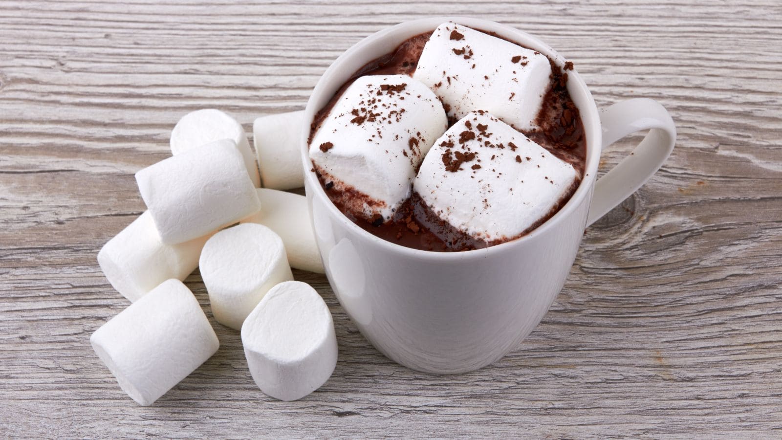 A white mug filled with hot chocolate topped with three large marshmallows sits on a wooden surface. Cocoa powder is sprinkled on top. Several plain marshmallows are piled beside the mug.