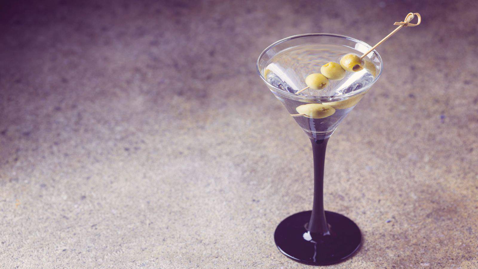 A cocktail glass with a black stem filled with a clear liquid, likely a martini, is placed on a textured surface. Three green olives are skewered on a toothpick and laid across the rim of the glass.