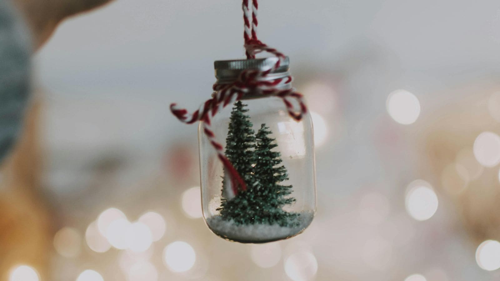 A small glass jar hangs from a red and white string. Inside the jar, there are miniature evergreen trees and a layer of white material resembling snow. Blurry lights in the background create a festive atmosphere.