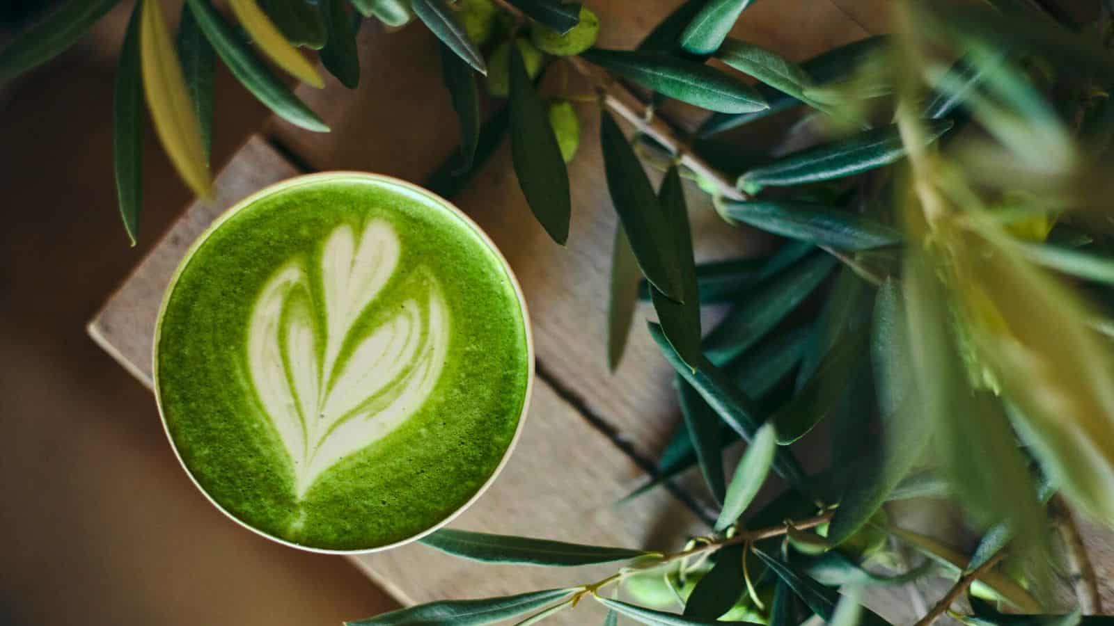 A cup of matcha latte with a white leaf pattern in the foam sits on a wooden surface. Green leaves surround the cup, creating a natural, soothing atmosphere.