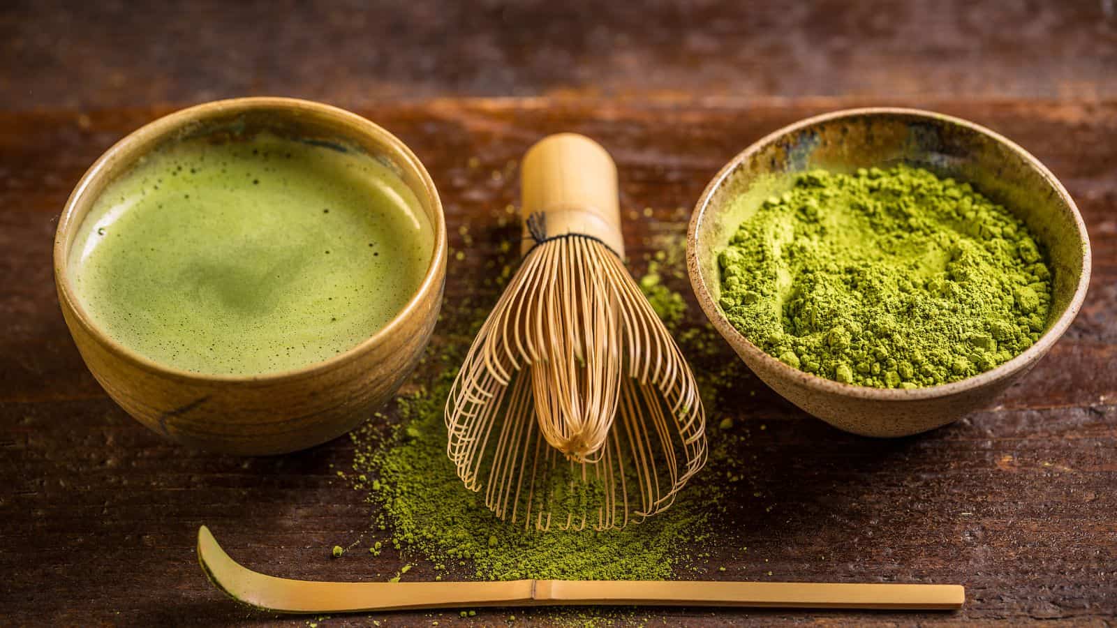 A wooden table with three items: a bowl of prepared matcha tea, a bamboo whisk, and a bowl of matcha powder. A bamboo scoop is placed in front, and some matcha powder is scattered on the table.