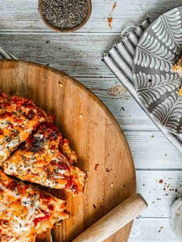 A wooden board with sliced pizza, topped with herbs and cheese, on a white wooden table. A striped cloth napkin is underneath one corner of the board. A small plate with a single pizza slice and pepper flakes in a jar are on the table.