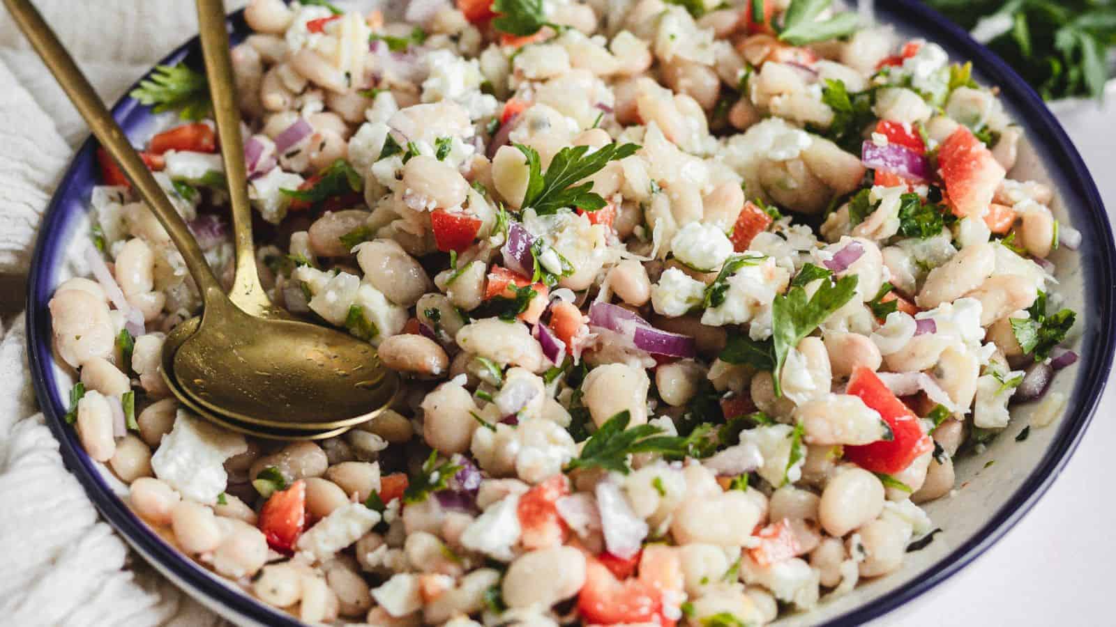 A close-up of a bowl filled with a white bean salad. The salad includes diced tomatoes, purple onions, chopped parsley, and crumbled feta cheese. Two golden spoons rest on the side of the bowl.
