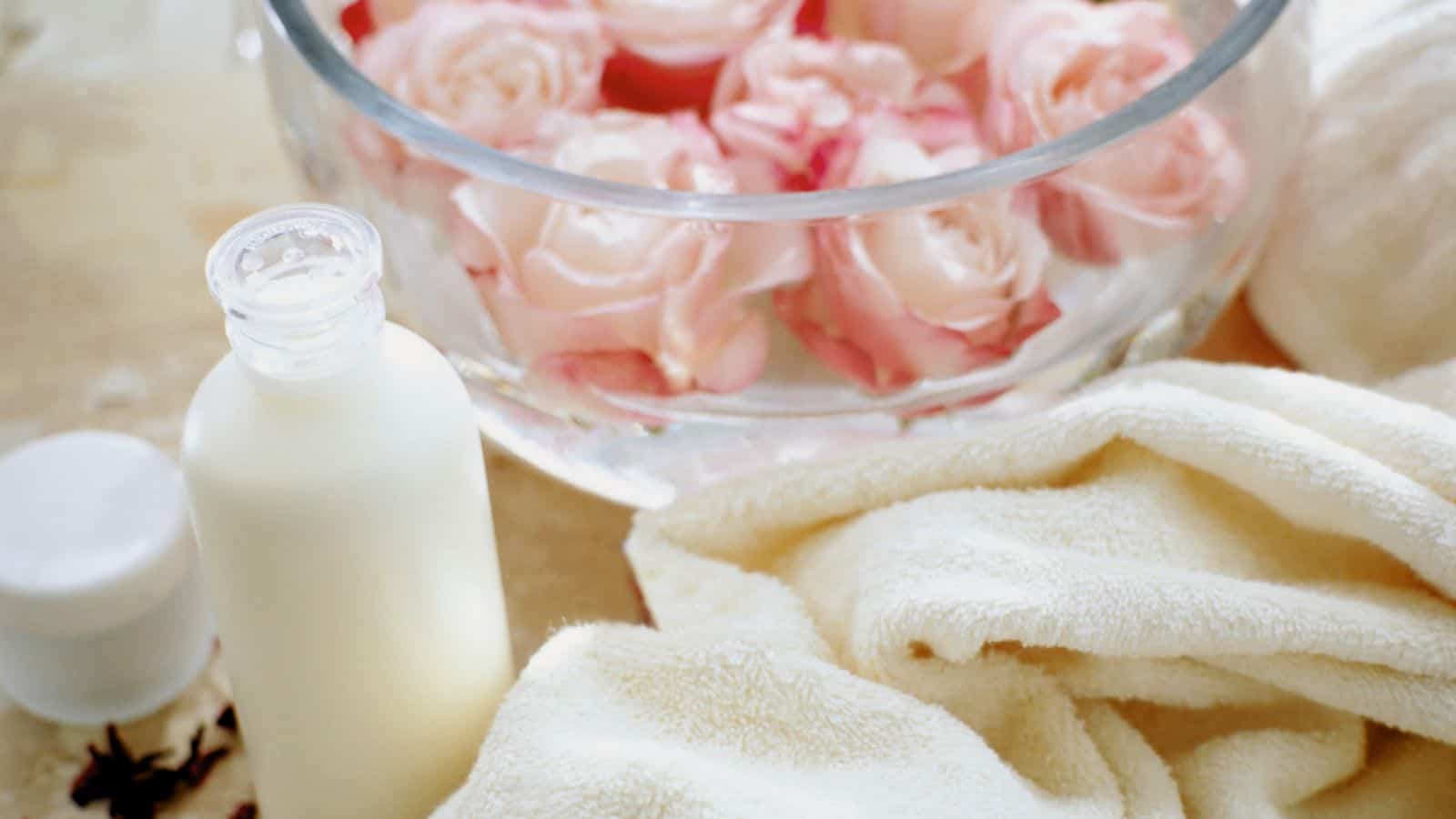 A glass bowl filled with pink roses is placed on a surface. Next to it, there is a white bottle with a cap and a small white jar. Cream-colored towels are folded nearby, suggesting a spa or relaxation setting.