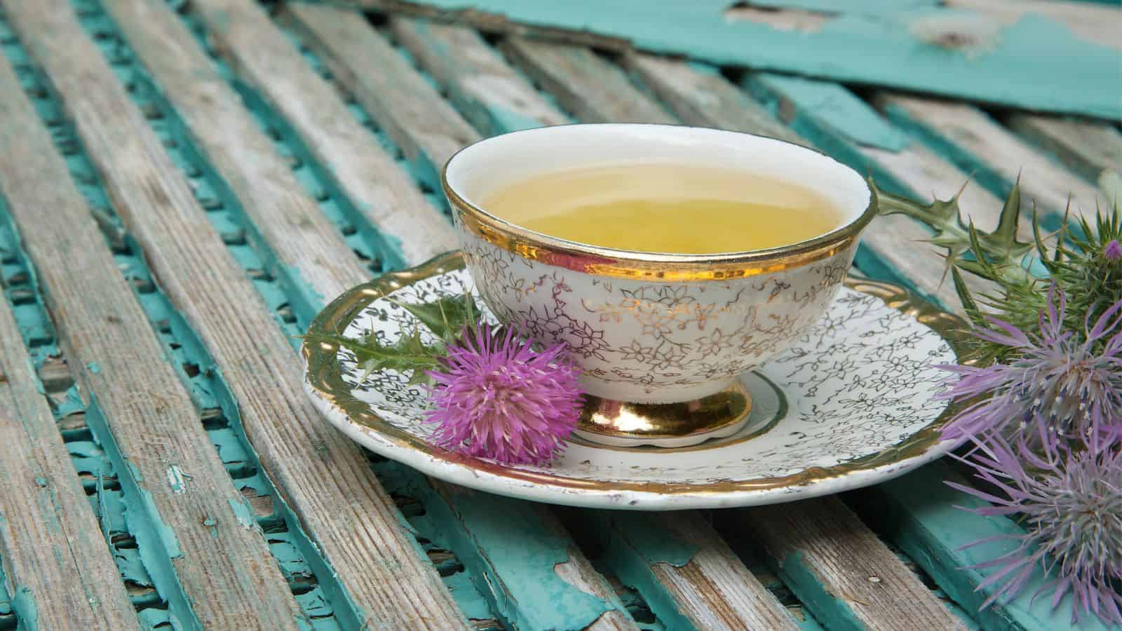 A floral-patterned teacup filled with light green herbal tea sits on a matching saucer. It is placed on a weathered turquoise wooden surface. Nearby, there are thistle flowers adding a touch of purple to the scene.