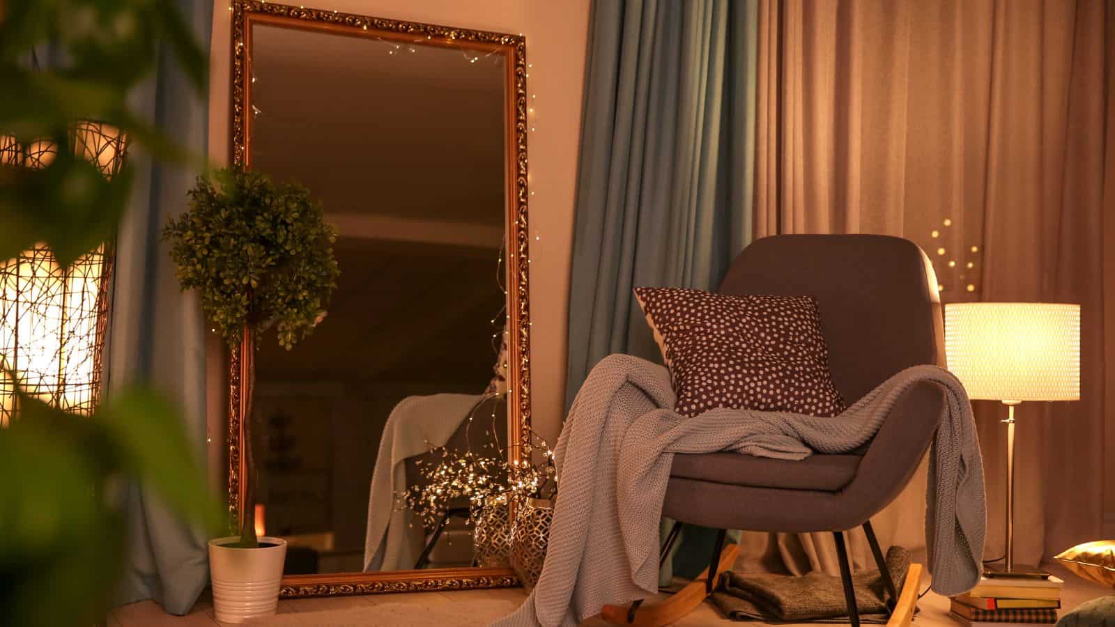 A cozy room with a large ornate mirror, a gray armchair draped with a blue blanket and dotted pillow. A potted plant sits beside the chair. A lit table lamp with a shade is nearby, and softly colored curtains are in the background.