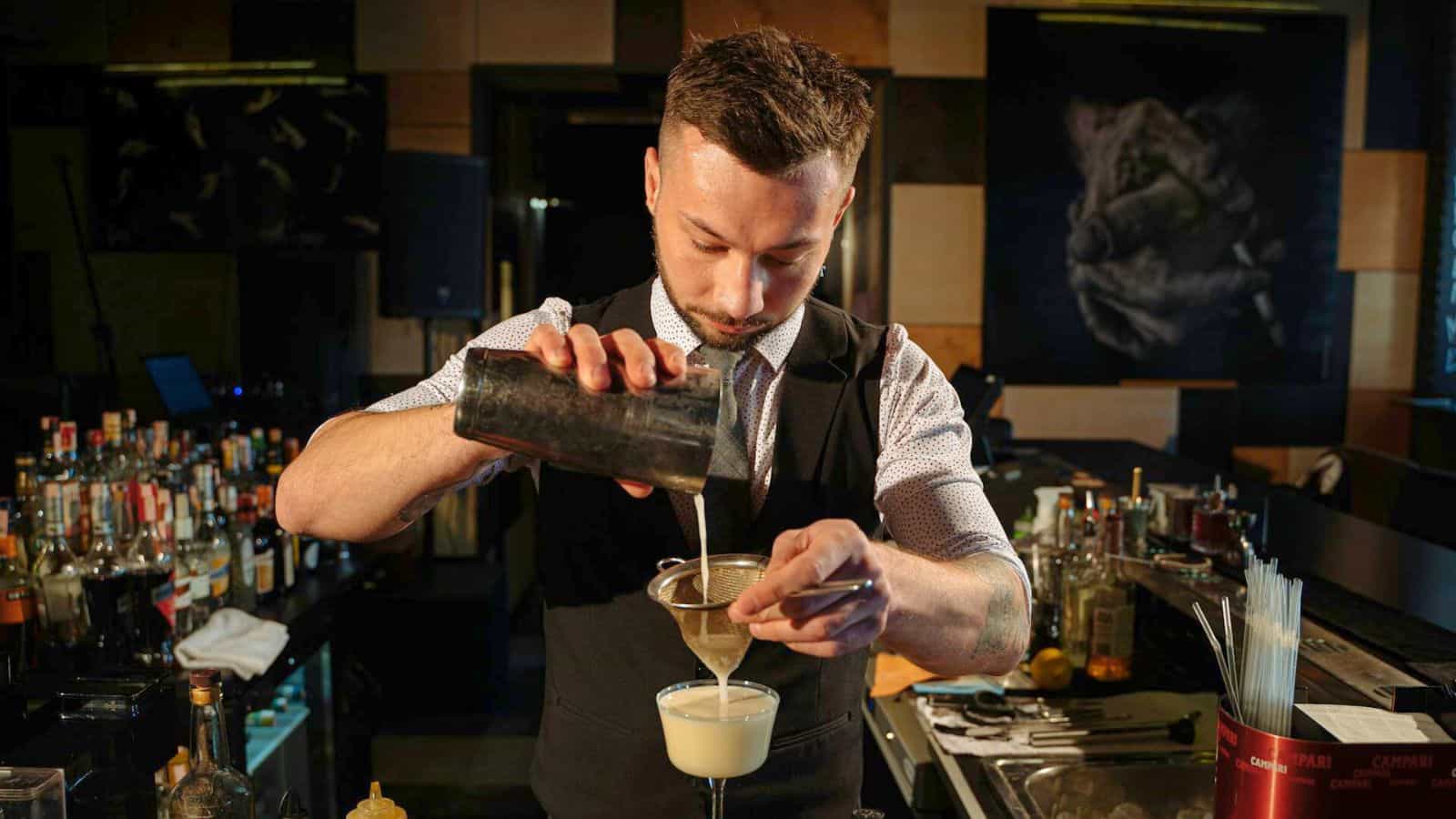 A bartender is pouring a drink through a strainer into a glass at a dimly lit bar. The bar is stocked with various bottles in the background, and cocktail-making tools are visible on the counter.