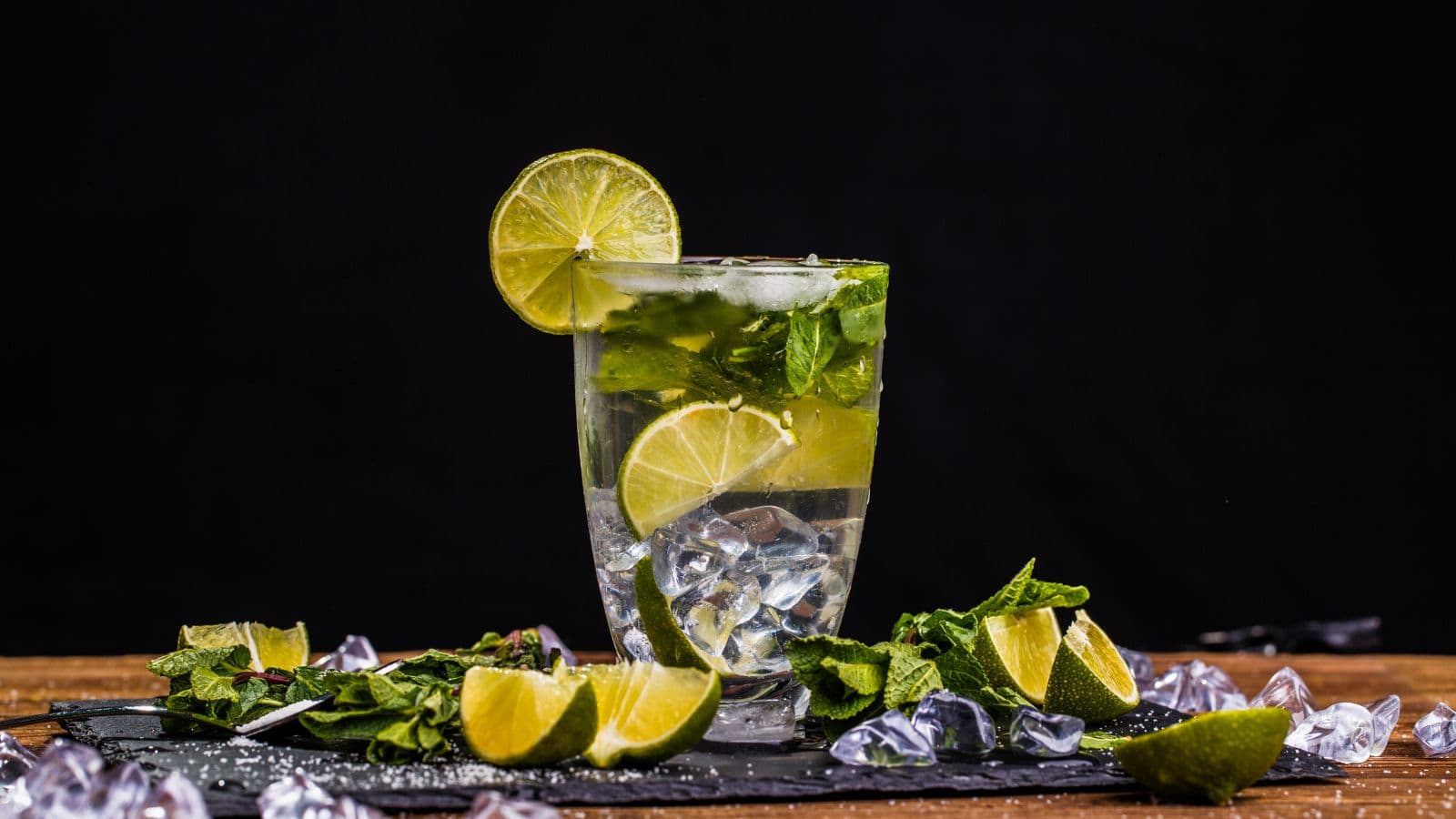 A tall glass of mojito filled with ice, lime slices, and mint leaves. The drink is garnished with a lime wheel on the rim. Additional lime wedges, mint, and ice cubes are scattered on a dark surface against a black background.