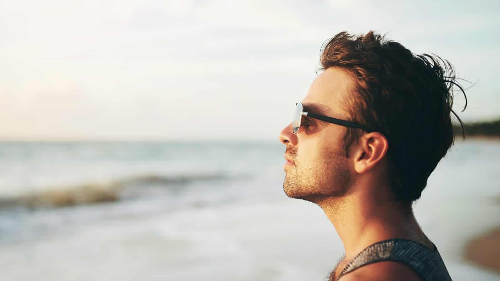 A person wearing sunglasses and a sleeveless shirt is standing on a beach, looking towards the ocean. The sky is clear and the waves are visible in the background.