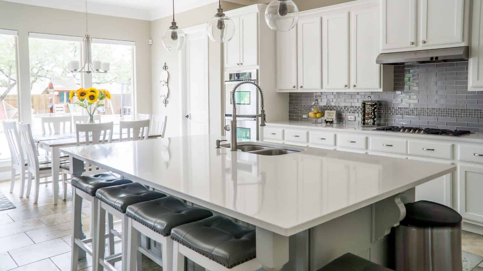 A modern kitchen with white cabinets and a large island with a white countertop. Three pendant lights hang above the island, which has gray cushioned stools. A dining table with sunflowers is near a window. Stainless steel appliances are visible.