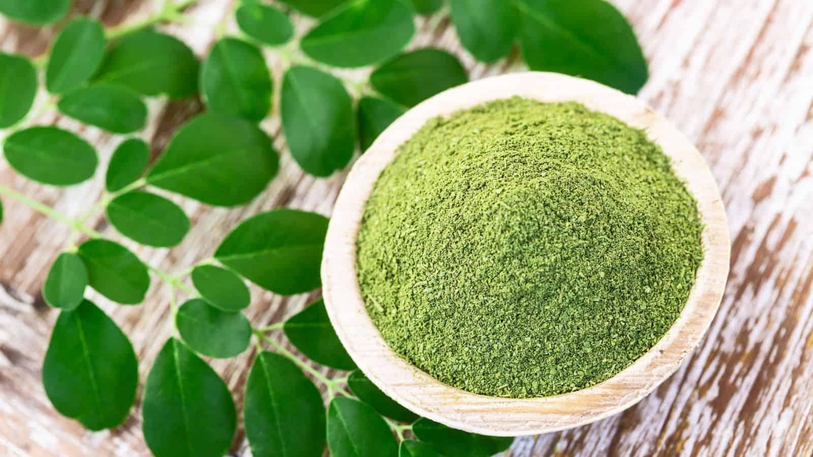 A wooden bowl filled with green powder, likely moringa, is placed on a wooden surface. Fresh green leaves are spread around the bowl. The background is textured, adding a natural feel to the composition.