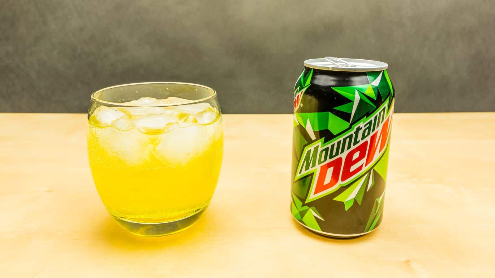 A can of Mountain Dew is placed on a wooden surface next to a glass filled with ice and a yellow liquid. The background is a neutral gray.