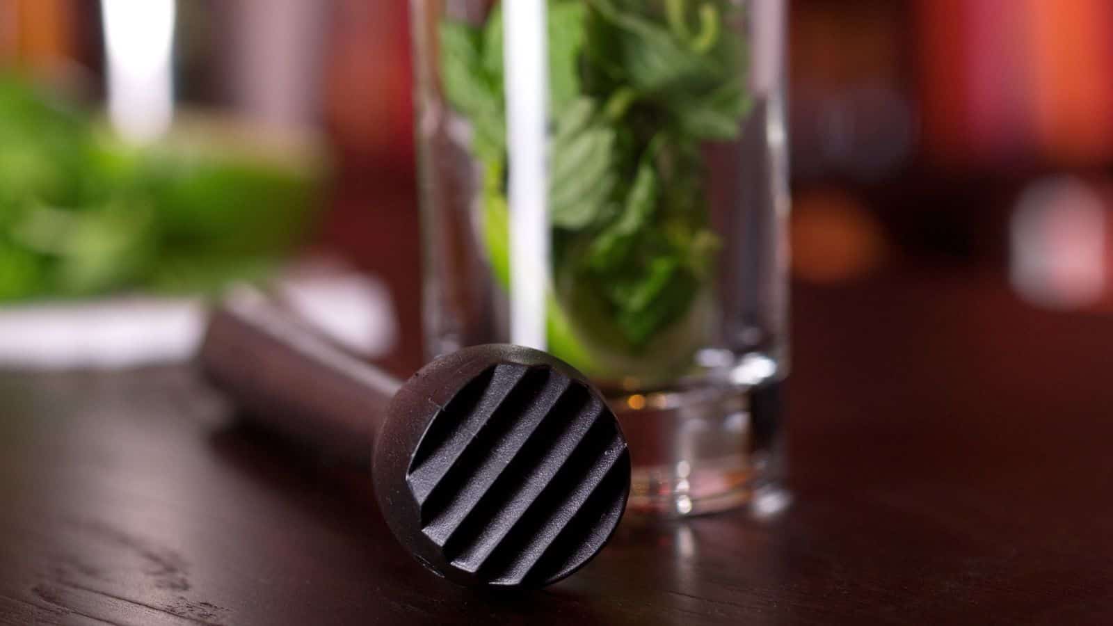 A close-up of a muddler on a dark wooden table, next to a glass filled with fresh mint leaves.