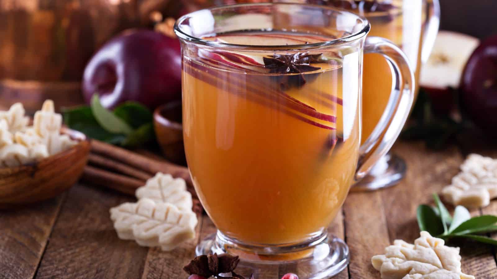 A glass mug filled with apple cider, garnished with cinnamon sticks and star anise, sits on a wooden table. Surrounding it are small leaf-shaped cookies, red apples, and cinnamon sticks, creating an autumnal theme.