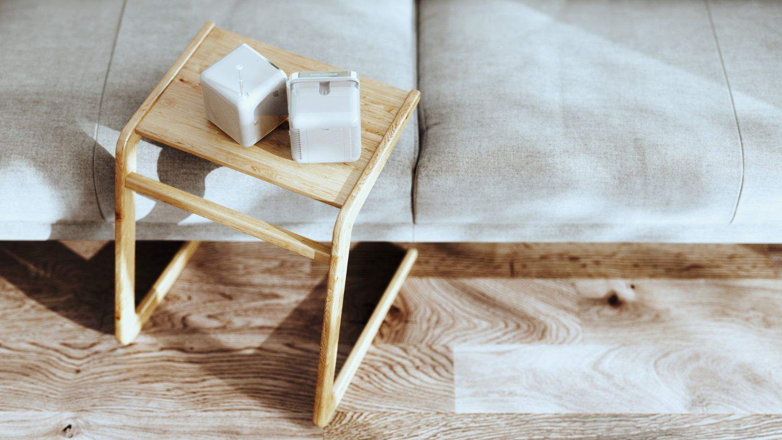 A small wooden table is placed beside a gray cushioned bench. On the table, there are two white cube-shaped objects with handles, possibly small speakers or containers. The scene is lit by natural sunlight, casting shadows on a wooden floor.