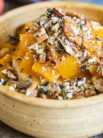 A ceramic bowl contains chopped peaches topped with crushed pecans and herbs. Nearby, there are whole pecans and part of a peach on a wooden surface. A gray cloth is partially visible to the right.