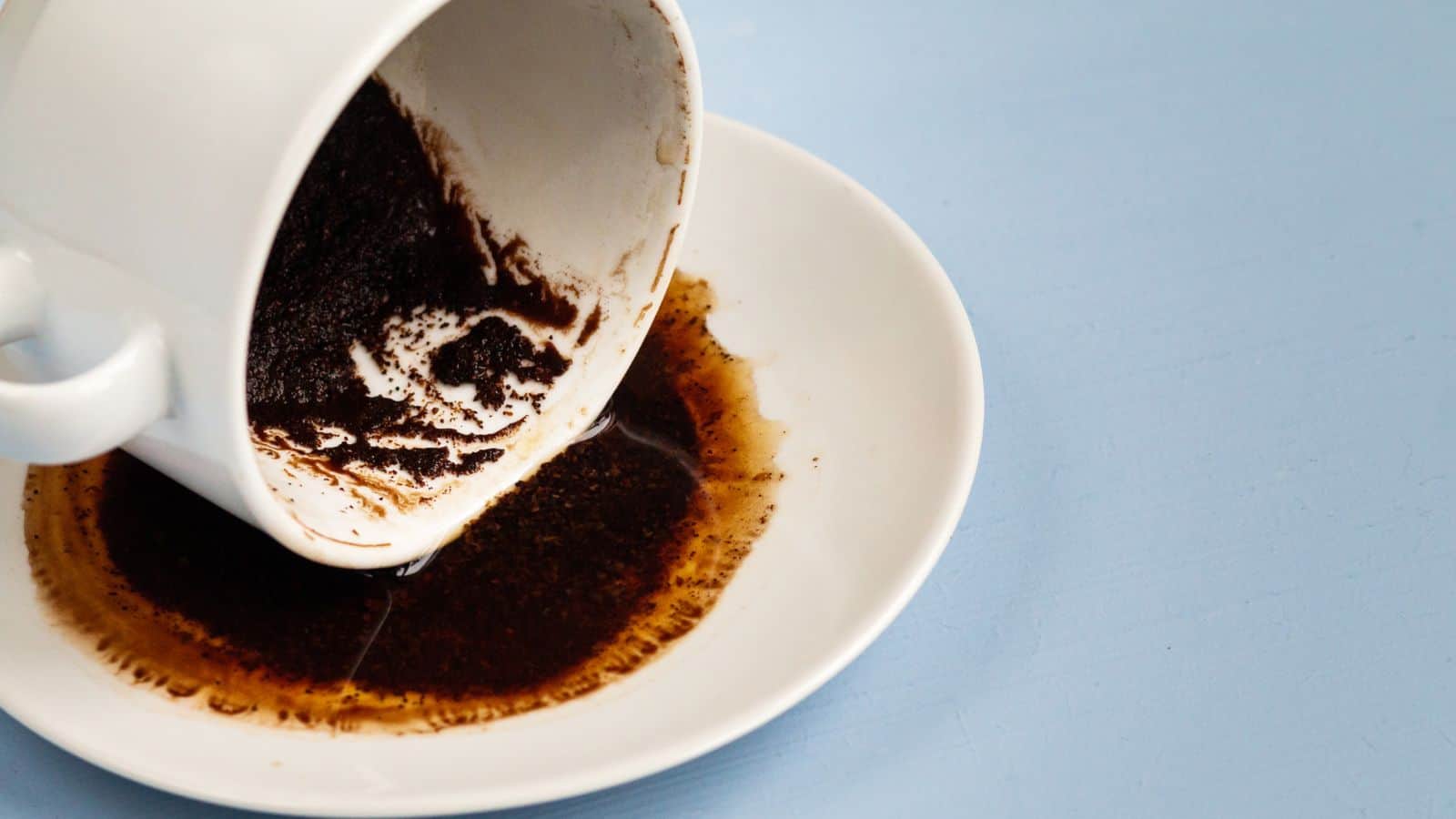 A white cup is tipped over on a white saucer, spilling dark brown coffee grounds and liquid onto the saucer. The background is a solid light blue color.