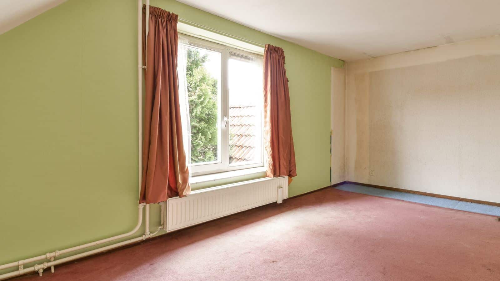 An empty room with a green wall and a window covered by red curtains. The floor is carpeted in a reddish color. White radiator and exposed pipes are beneath the window. Light enters through the window, illuminating the space.