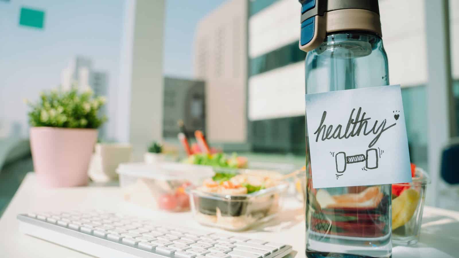 A desk with a clear water bottle labeled "healthy," a white keyboard, and containers of salads and snacks. A small potted plant is in the background, with a cityscape visible through a window.