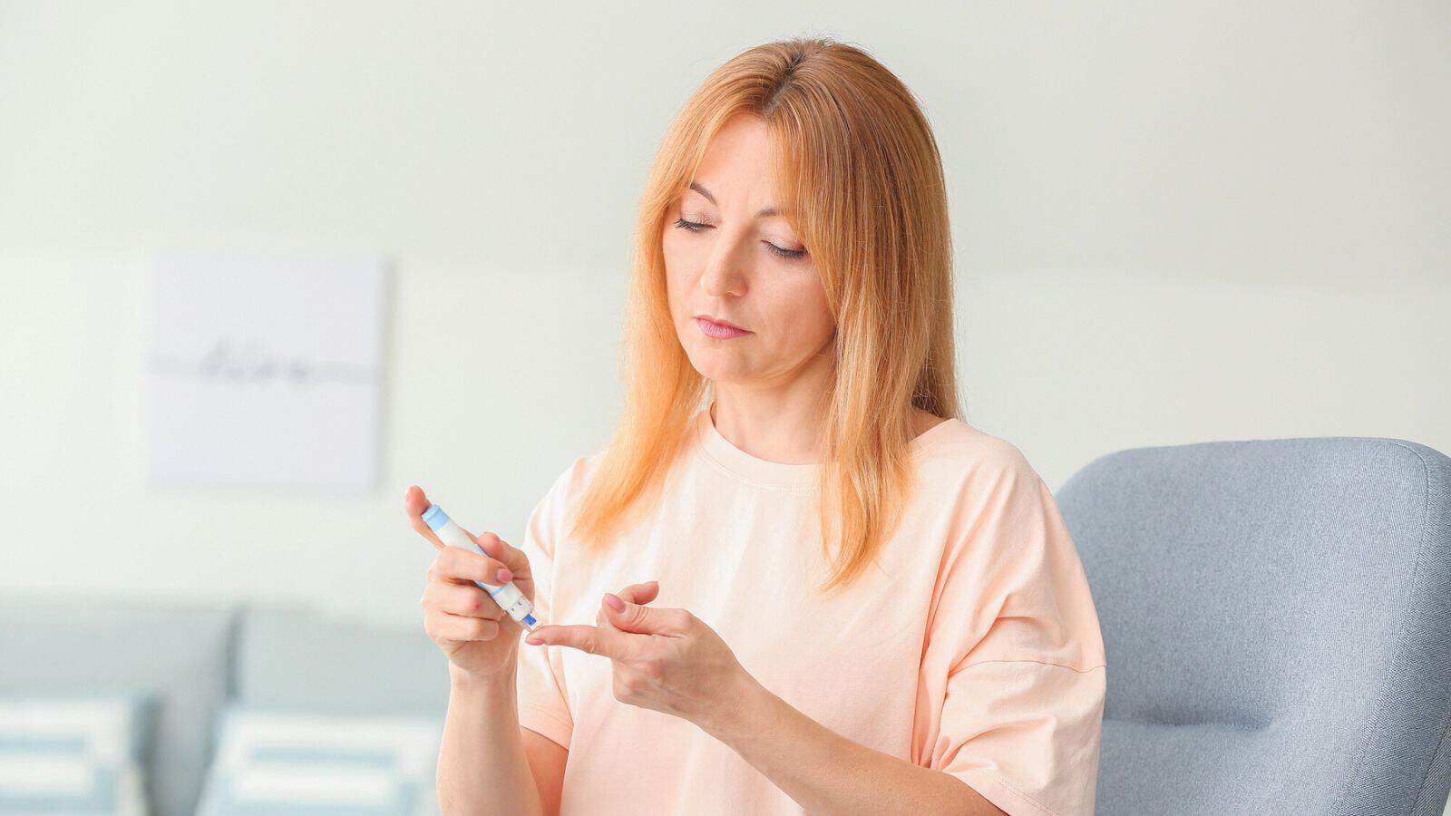 A person with long hair is sitting in a chair, using a lancet device to check their blood sugar. The background is softly blurred, with hints of light-colored furniture. The individual is focused on their fingertip.