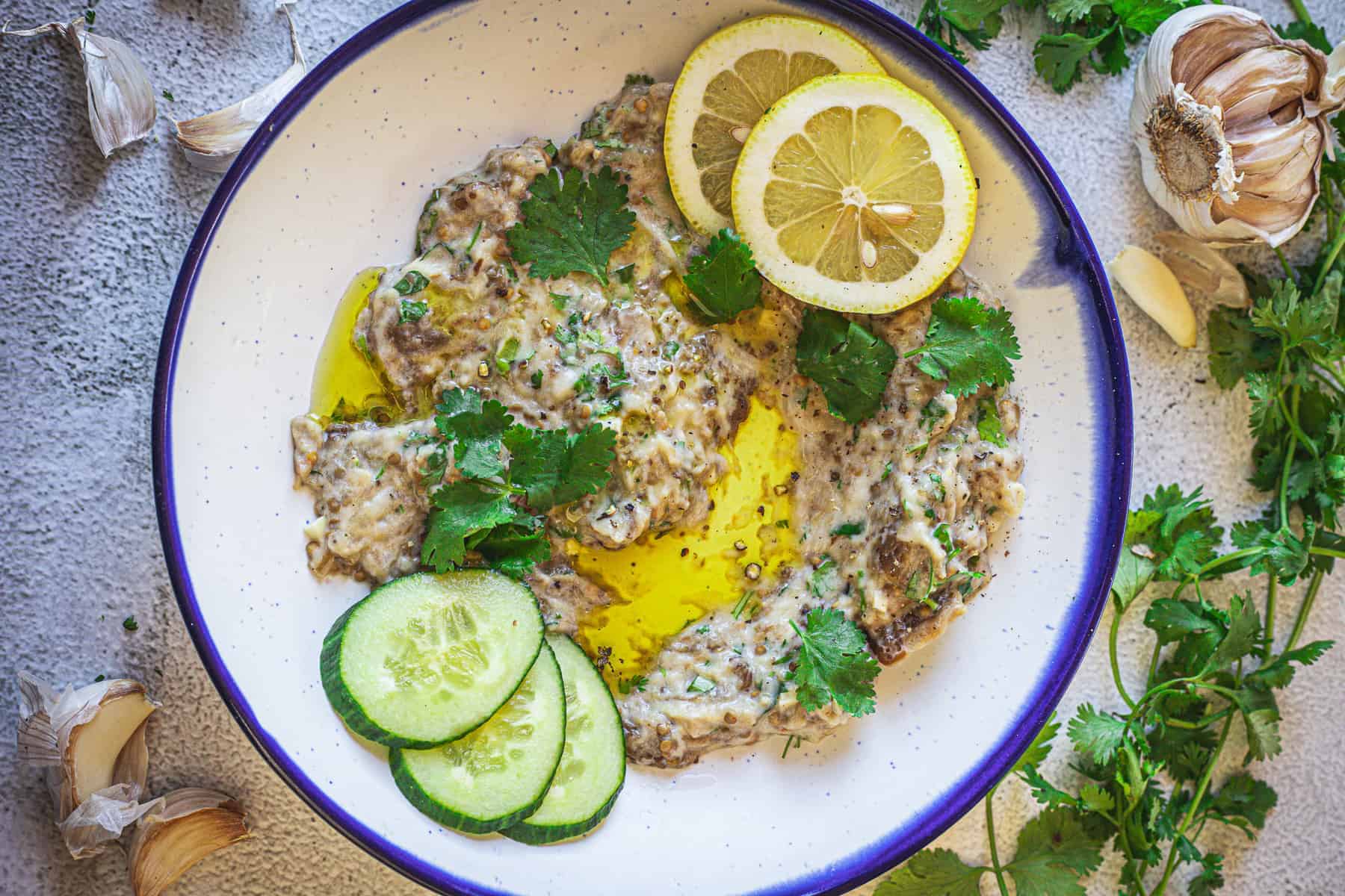 A plate of baba ganoush garnished with cilantro leaves, lemon slices, and cucumber slices. Drizzled with olive oil, it is surrounded by garlic cloves and cilantro sprigs on a textured surface.