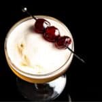A cocktail in a glass with a frothy top, garnished with three cherries skewered on a metal stick, set against a black background.