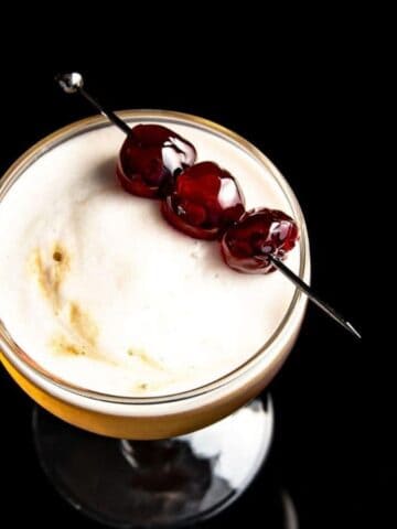 A cocktail in a glass with a frothy top, garnished with three dark cherries on a skewer, set against a black background.