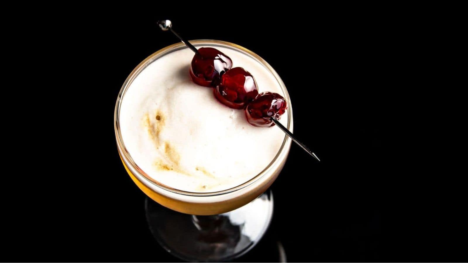 A cocktail in a glass with a frothy top, garnished with three cherries skewered on a metal stick, set against a black background.