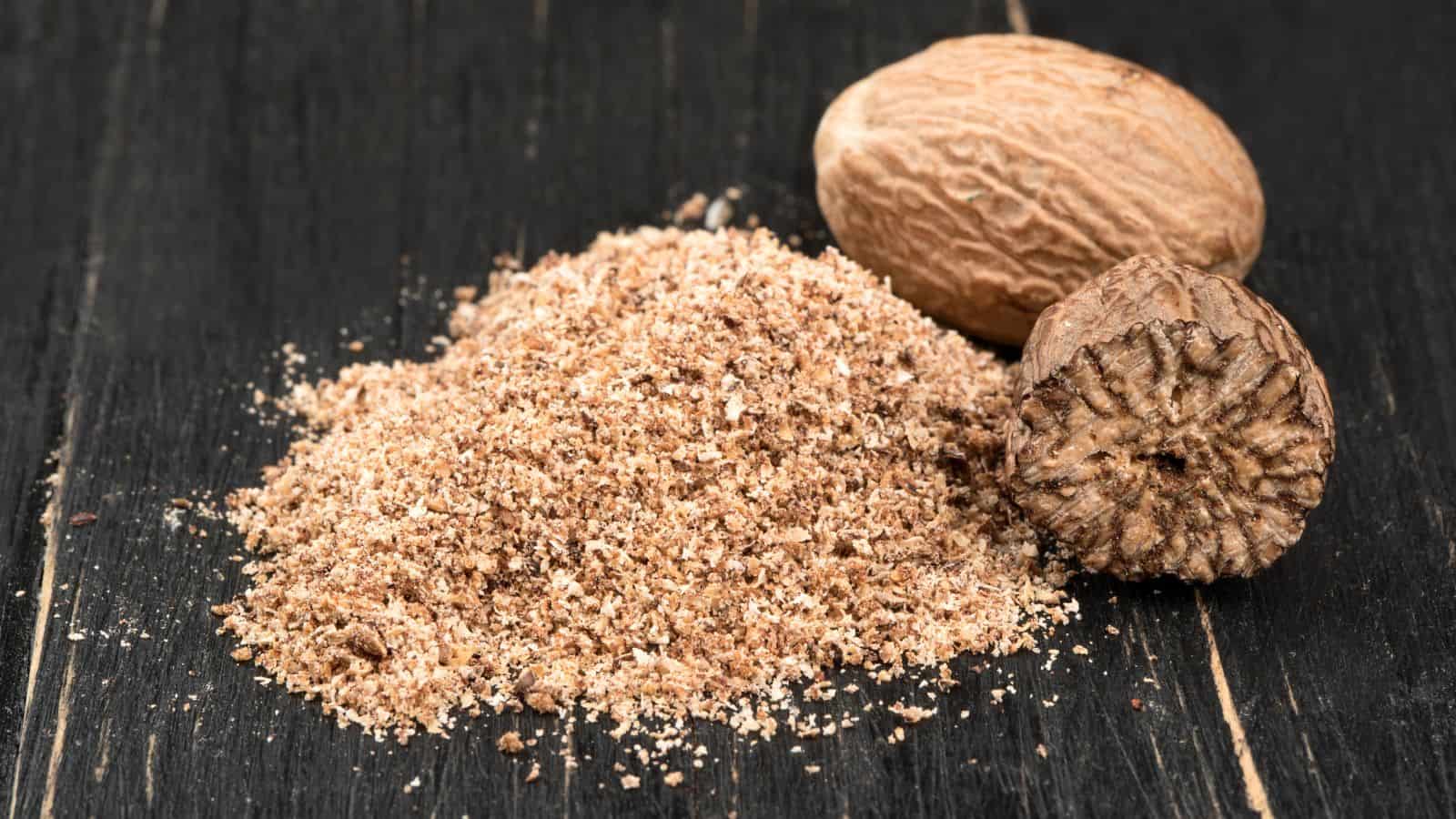 Whole and ground nutmeg on a dark wooden surface. The ground nutmeg is in a small pile beside two whole nutmegs, one partially cut open to reveal its interior texture.