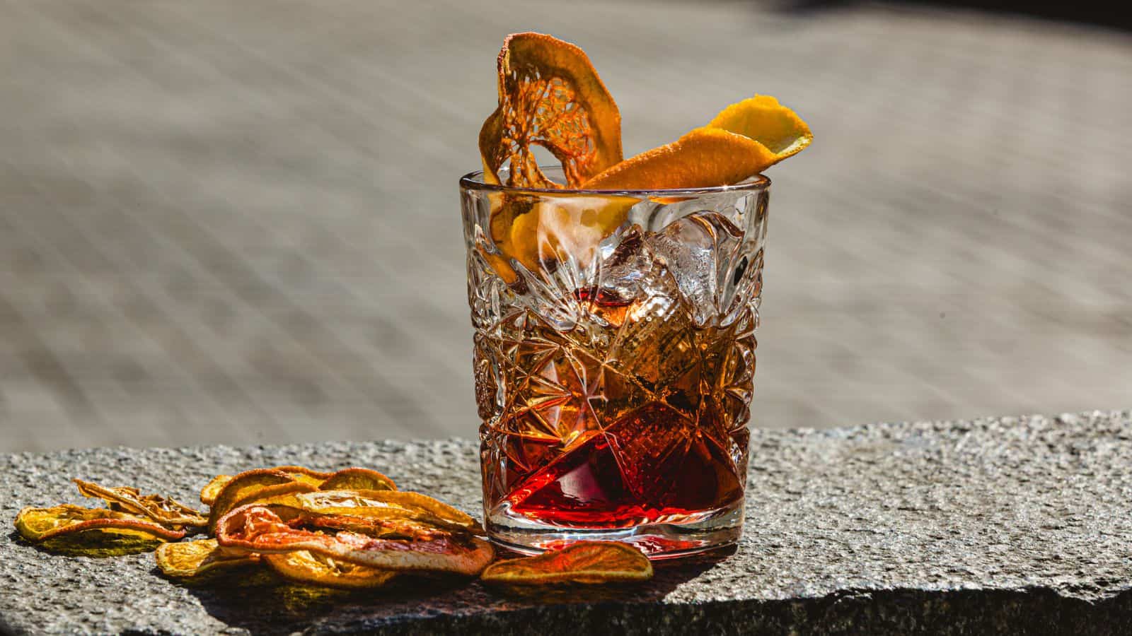 A crystal glass with a red cocktail sits on a stone surface, featuring ice and garnished with dried orange slices. Additional dried orange slices are placed beside the glass. The background is blurred.