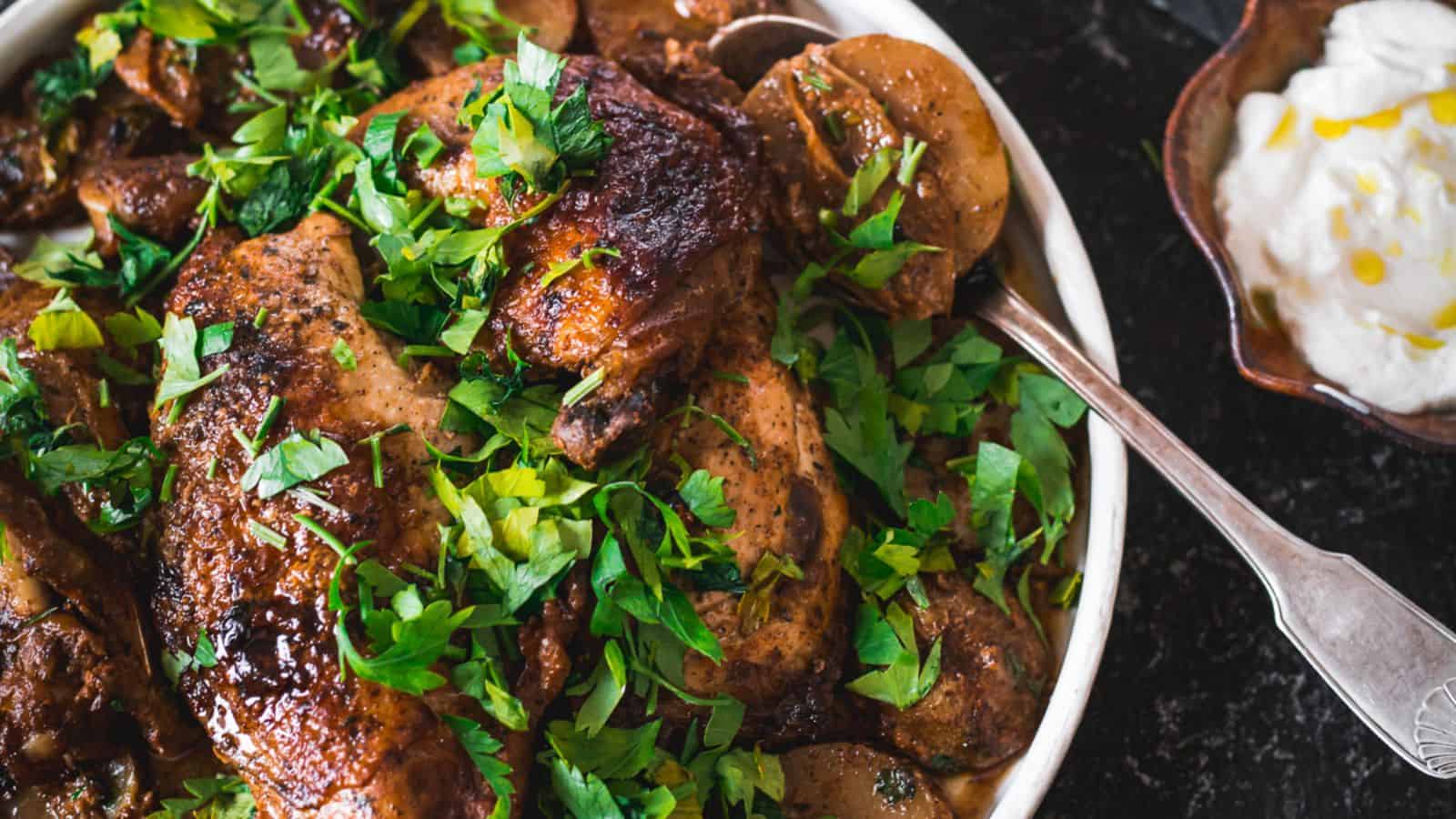 A roasted chicken dish garnished with fresh parsley on a white plate. Sliced potatoes surround the chicken. A side bowl with a white creamy sauce is visible on the right. A serving spoon rests on the plate.