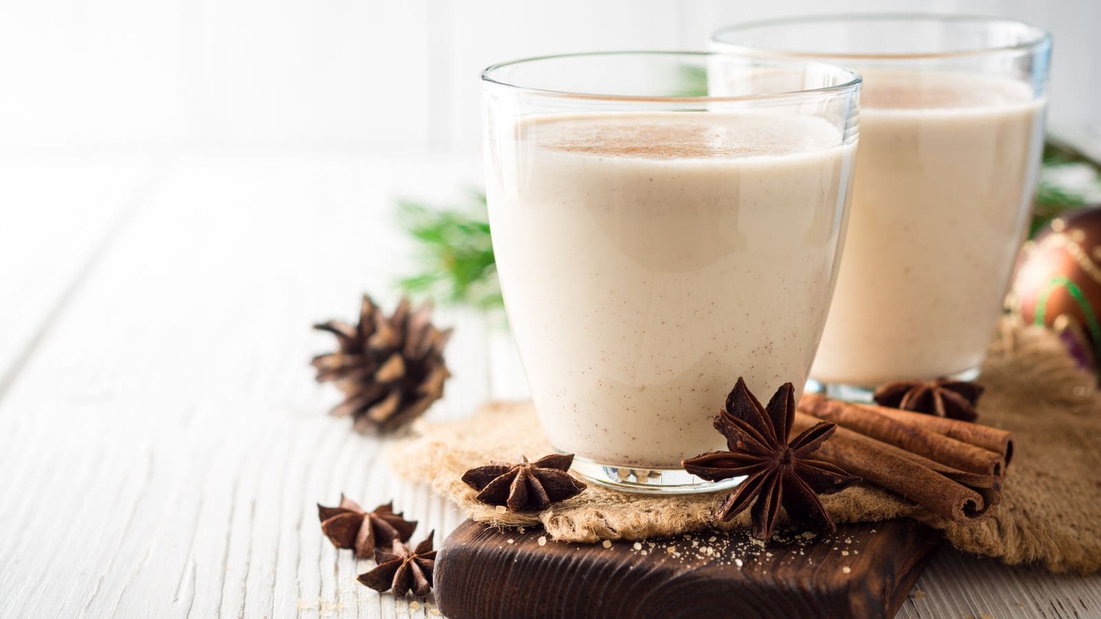 An image of two glasses of eggnog with pinecones and Chinese star anise scattered on the surface.