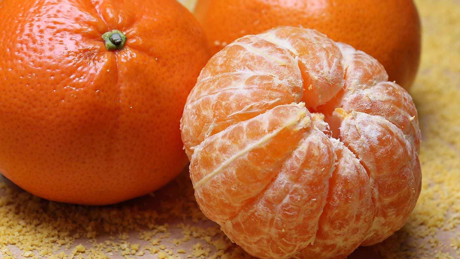 A close-up of a whole unpeeled orange and a peeled orange next to each other on a light surface. In the background, there is another unpeeled orange. Crumbs are scattered around the fruit.