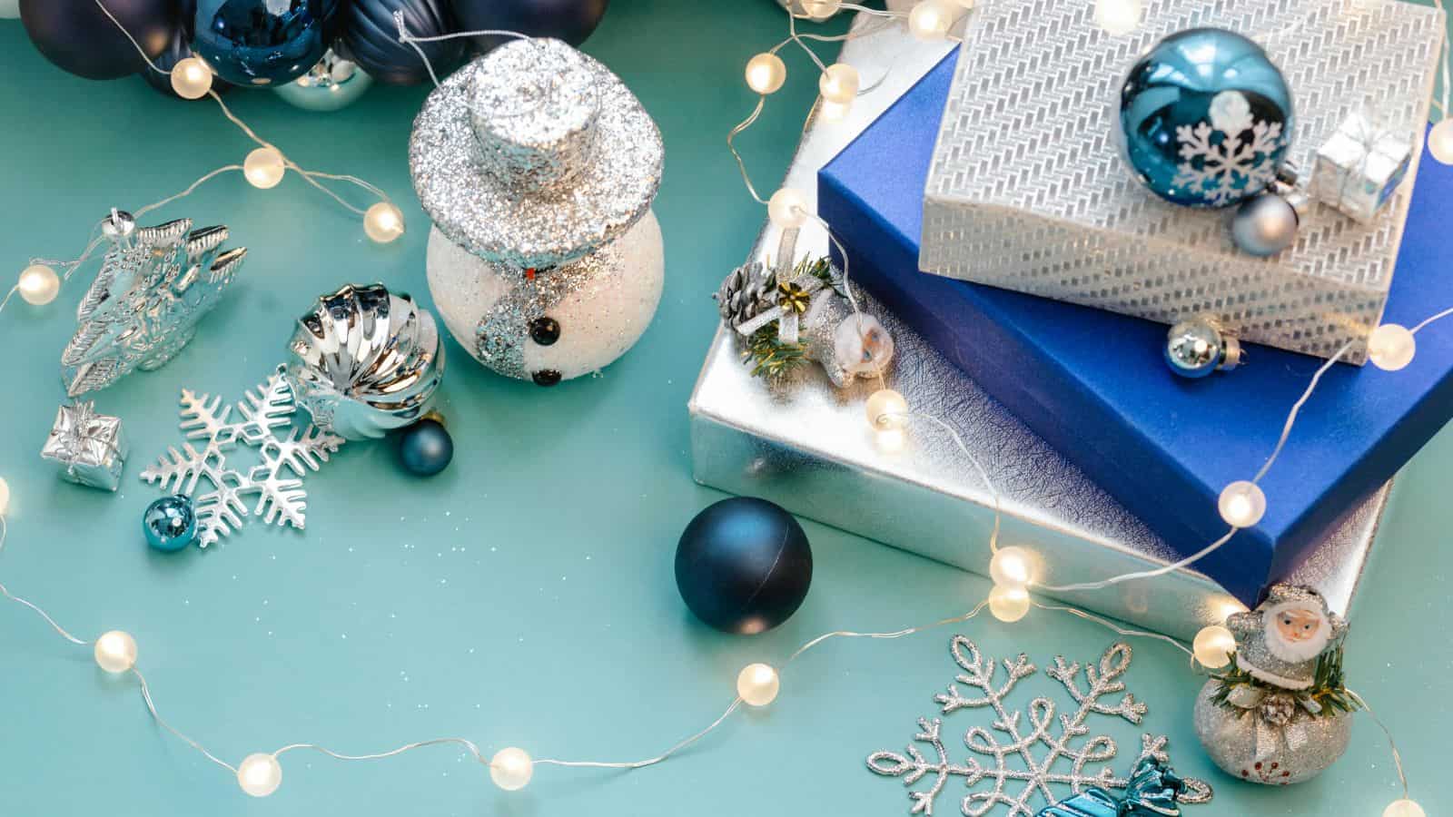 A festive scene with blue and silver gift boxes, a glittery snowman ornament, blue and silver baubles, and a string of lights. Snowflake decorations and a miniature snowman add to the holiday theme on a teal background.