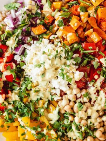 A colorful salad on a white plate featuring chickpeas, diced red and yellow bell peppers, chopped red onions, roasted sweet potatoes, fresh herbs, and a creamy dressing. A spoon is placed on the side. The plate is on a marble surface.